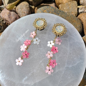 May include: A pair of gold and white ear gauges with a dangling chain of pink and white flowers. The gauges have a sparkly iridescent center.