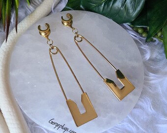 May include: A pair of gold-coloured ear gauges, each featuring a safety pin design. The gauges have a curved top and a rectangular base with a cutout. The website "Gorgeplugs.com" is visible in the image.