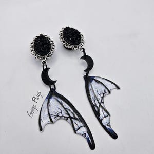 May include: A pair of black and silver earrings with a gothic theme. The earrings feature black druzy plugs with a silver filigree setting, connected to black crescent moons and wing-shaped dangles with a tree branch design. The text "Gorge Plugs" is visible.