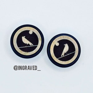 May include: Two round wooden earrings with a black background and a gold rim. Each earring features a white silhouette of a bird perched on a wire with a moon in the background. The text "@INGRAVED_" is visible below the earrings.