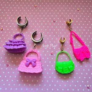 Peut inclure: Quatre écarteurs d'oreille colorés avec des breloques en forme de sac à main. Les écarteurs sont argentés et dorés, avec des breloques violettes, roses, vertes et rose vif. Le sac à main rose a un nœud. Le texte "Gorge Plugs" est visible.