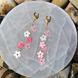 May include: A pair of gold-toned ear cuffs with a dangling chain of pink and white flower charms. The charms are shaped like cherry blossoms and are arranged in a cascading pattern. The ear cuffs are a popular accessory for women.