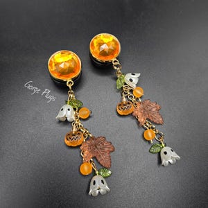 May include: A pair of ear plugs with faceted orange circular tops and gold-tone chains. Each chain features dangling charms including white ghost-shaped beads, orange pumpkin-shaped beads, brown maple leaf charms, and small green leaf accents.