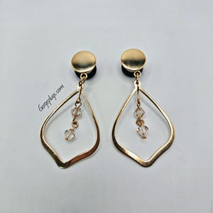 May include: A pair of gold dangle earrings with a teardrop-shaped design. Each earring features a small round gold disc at the top and a dangling chain with three clear crystal beads.