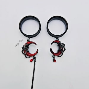 May include: Pair of black ear gauges with red crescent moon charms and dangling black chains. The moon charms have a dark, gothic aesthetic with a drop-shaped red gem. The text "Gorge Plugs" is visible.