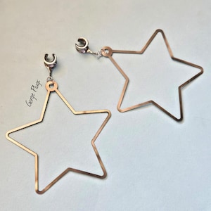 May include: A pair of silver star-shaped earrings with a clip-on design. The earrings are large and have a simple, modern style.