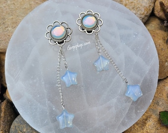 May include: A pair of iridescent opal plugs with silver flower accents and dangling star charms. The plugs are on a white surface with the text "Gorgeplugs.com".