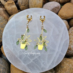 May include: A pair of gold hanging plant earrings with green leaves. The earrings are made of metal and have a hook closure. The earrings are on a white background.
