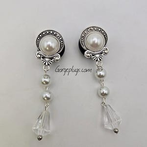 May include: A pair of silver ear gauges with a pearl and crystal dangle. The gauges have a decorative design with a pearl in the center. The dangle features a crystal at the bottom and three white pearls.