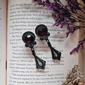 May include: A pair of black earrings with a gothic design. Each earring features a large, dark red gemstone and intricate metalwork. The earrings are displayed on a book with text about a fictional character named Jagger.