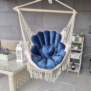 May include: A white macrame hammock chair with a blue velvet cushion and a heart-shaped pillow. The chair is hanging from a wooden beam and has a fringe trim.