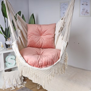 Peut inclure: Un fauteuil hamac en macramé blanc avec un coussin en velours rose. Le hamac est suspendu au plafond et possède une bordure en franges.