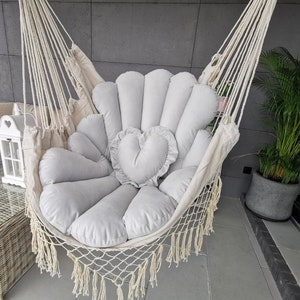 Peut inclure: Un fauteuil hamac blanc avec un coussin en forme de cœur gris et un coussin en forme de fleur gris. Le hamac est suspendu au plafond et est fait de corde.