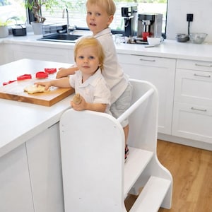 Op de afbeelding: Een witte houten keukentrap met twee kinderen die erop staan. Eén kind houdt een deegroller vast en de ander werkt met deeg en rode koekjesvormen op een houten snijplank.