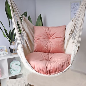 May include: A white hammock chair with a pink cushion and a pink pillow. The hammock is hanging from the ceiling and is in a room with white walls and a white shelf.
