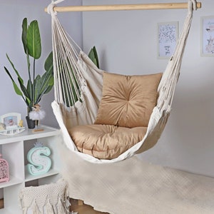 May include: A white hammock chair with a beige cushion. The hammock is hanging from a wooden bar and is suspended in mid-air. The chair is in a room with white walls and a white rug.