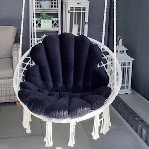 Peut inclure: Un fauteuil suspendu en macramé blanc avec un coussin noir en forme de coquillage. La chaise est suspendue par d'épaisses cordes blanches et comporte des franges décoratives en bas. L'arrière-plan comprend un patio aux tons neutres et des étagères décoratives.