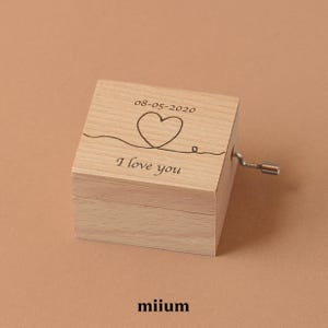 May include: A light-coloured wooden music box with a hand crank. The top is engraved with a heart, the date 08-05-2020, and the words "I love you". The brand name "miium" is at the bottom. The box is made of light-coloured wood.