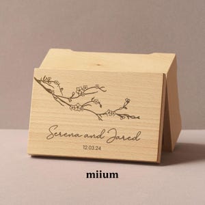 Può includere: Una scatola di legno color chiaro con coperchio incernierato. La parte anteriore della scatola presenta un delicato design a ramo floreale e i nomi "Serena and Jared" con la data "12.03.24" incisa sotto. La parola "miium" è in basso.