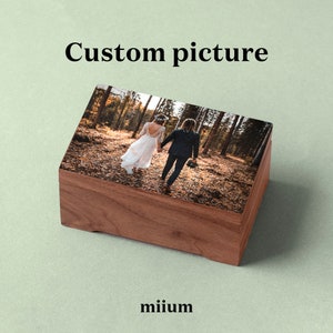 May include: A wooden box with a hinged lid, featuring a printed image of a couple walking in a forest. The box is made of dark wood with a smooth finish.