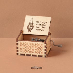 May include: A wooden music box with a crank and etched floral design. The lid features a drawing of a gnome and the French text "les temps sont durs pour les rêveurs". The brand name "miium" is at the bottom.