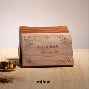 Puede incluir: Caja de música de madera con la palabra "COLDPLAY" grabada en la parte delantera, junto con la fecha "Coimbra 18 Maio 2023" y el nombre "Ana Mauricio". La caja presenta diseños celestiales y el nombre de la marca "miium".