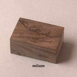 May include: A rectangular wooden box with a hinged lid. The lid is engraved with a floral branch design and the names "Serena and Jared" along with the date "12.03.24". The box is a warm brown color and has a smooth finish. The word "miium" is at the bottom.