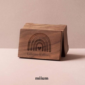 Puede incluir: Un bloque de madera marrón con un diseño de arcoíris tallado y el nombre "Katherine Williams" grabado debajo. El bloque tiene un corazón en el centro del arcoíris. La palabra "miium" está en la parte inferior.