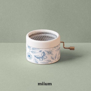 May include: White music box with a blue and white floral design featuring butterflies and bees. The music box has a metal winding key and a mesh screen on the top.