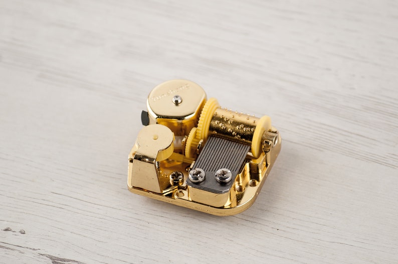 Puede incluir: Un mecanismo de caja de m&uacute;sica de color dorado con una llave de cuerda y una peque&ntilde;a placa de metal con agujeros. El mecanismo est&aacute; sobre una superficie de madera blanca.