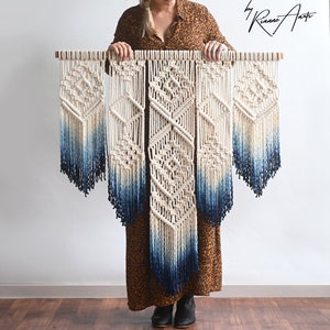 May include: A woman holds a large macrame wall hanging with a blue ombre design. The wall hanging is made of natural cotton cord and has a geometric pattern. The text "CONTEMPORARY fiber art by Rianne Arts" is visible in the top right corner of the image.