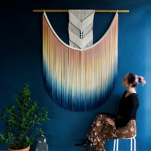 May include: A large macrame wall hanging with a gradient of peach, yellow, blue, and navy colours. The macrame is hanging on a dark blue wall. The macrame is made with a variety of knots and has a fringe at the bottom. The macrame is a popular home decor item.