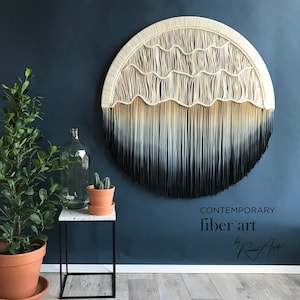 May include: A large, circular macrame wall hanging with a white and black ombre design. The hanging is made of natural fiber rope and has a fringe border. The wall hanging is displayed against a dark blue wall with the text "CONTEMPORARY fiber art by Riana Art" below it.