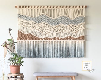 May include: A large wall hanging made of macrame with a woven design in shades of white, brown, and blue. The hanging has a fringe border and is mounted on a wooden dowel. The hanging is displayed on a white wall with a gray bench in front of it. The text "Teddy and Wool" is visible in the lower right corner of the image.