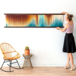 Op de afbeelding: Een vrouw hangt een kleurrijk wandtapijt van garen aan een witte muur. Het wandtapijt heeft een verloop van oranje, geel, blauw en groene kleuren. Het wandtapijt is ingelijst met een donkerhouten lijst.