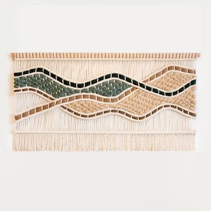 May include: A large, rectangular macrame wall hanging with a natural, neutral color palette. The design features undulating waves in shades of cream, green, tan, and brown, with a fringed bottom edge. The piece is mounted on a wooden dowel.