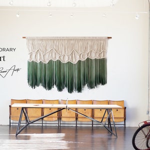 May include: Contemporary fiber art by Riana Art. A large wall hanging with a white and green ombre design. The hanging is made of yarn and has a wave-like pattern. The hanging is displayed in a white room with a wooden table and chairs.