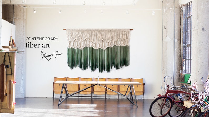 May include: Contemporary fiber art by Riana Art. A large wall hanging with a white and green ombre design. The hanging is made of yarn and has a wave-like pattern. The hanging is displayed in a white room with a wooden table and chairs.