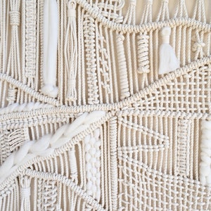Puede incluir: Primer plano de un tapiz de macram&eacute; hecho con cord&oacute;n de algod&oacute;n blanco natural. El tapiz presenta una variedad de nudos y patrones, creando un dise&ntilde;o texturizado e intrincado.
