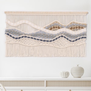 May include: A macrame wall hanging with a woven design featuring a wavy pattern in shades of beige, gray, and blue. The hanging has a fringe border and is mounted on a wooden dowel.