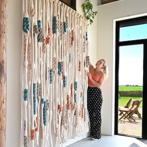 Op de afbeelding: Een grote wanddecoratie gemaakt van geweven touw in verschillende kleuren, waaronder wit, beige, teal en oranje. Het touw is geknoopt en gelust om een uniek en textuurrijk ontwerp te creëren.