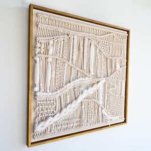 Puede incluir: Un gran tapiz de macram&eacute; blanco con un dise&ntilde;o geom&eacute;trico. El macram&eacute; est&aacute; enmarcado en un marco dorado.