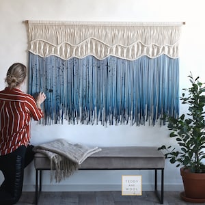 May include: A large wall hanging made of macrame with a blue ombre fringe. The fringe is made of yarn and is tied to a wooden dowel. The wall hanging is hanging on a white wall. The text "Teddy and Wool" is visible on a sign in the lower right corner of the image.