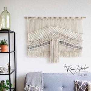 Op de afbeelding: Een grote, witte en grijze macramé wandhanger met een houten lat aan de bovenkant. De hanger heeft een golvend patroon met franjes aan de onderkant. De tekst "by Rianne Zuijderduin Teddy + Wool" is zichtbaar onderaan de afbeelding.