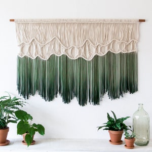 May include: A macrame wall hanging with a white and green ombre fringe. The hanging features a geometric pattern with a wave-like design.