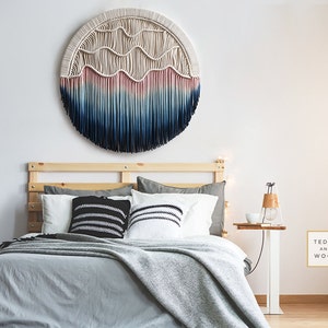 May include: A bedroom with a white wall, a wooden bed frame, and a large, round macrame wall hanging with blue, pink, and white yarn. The bed has a gray blanket and black and white patterned pillows. A small wooden side table with a lamp and a white cup is next to the bed. A framed print with the text "Teddy and Wool" is on the wall.