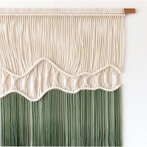 May include: A macrame wall hanging with a white top section and a green bottom section. The top section features a wave pattern and the bottom section is made of long strands of yarn.