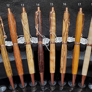 May include: Eight wooden pens with copper accents displayed on a black stand. The pens are made from different types of wood, including maple, walnut, and cherry. Each pen has a unique grain pattern and color.