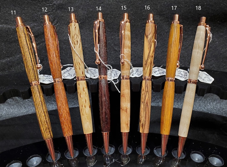 May include: Eight wooden pens with copper accents displayed on a black stand. The pens are made from different types of wood, including maple, walnut, and cherry. Each pen has a unique grain pattern and color.