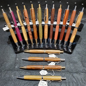 May include: A display of 14 wooden pens with gold accents and silver tips. The pens are arranged in a black stand with numbered slots. The pens are made from different types of wood, including walnut, maple, and cherry. The pens are all in a horizontal position, with the tips facing the right side of the image. The pens are all in focus, and the background is blurred.